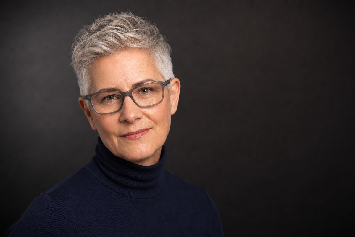 Professional studio headshot in a navy turtleneck and glasses against a dark background.